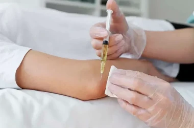 A gloved hand holds a syringe with yellow liquid while another gloved hand presses gauze on a person’s forearm.