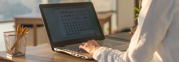 Close-up of a person using a laptop on a desk, with a keyboard, screen icons visible, and a pencil holder nearby.