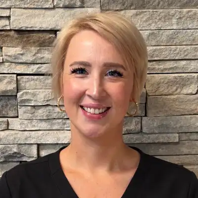 Professional staff headshot of a smiling person with short blonde hair wearing a black top, standing in front of a gray stacked stone wall background.