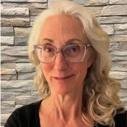Professional headshot of a person with long gray hair and glasses wearing a black top, photographed in front of a gray stacked stone wall background.
